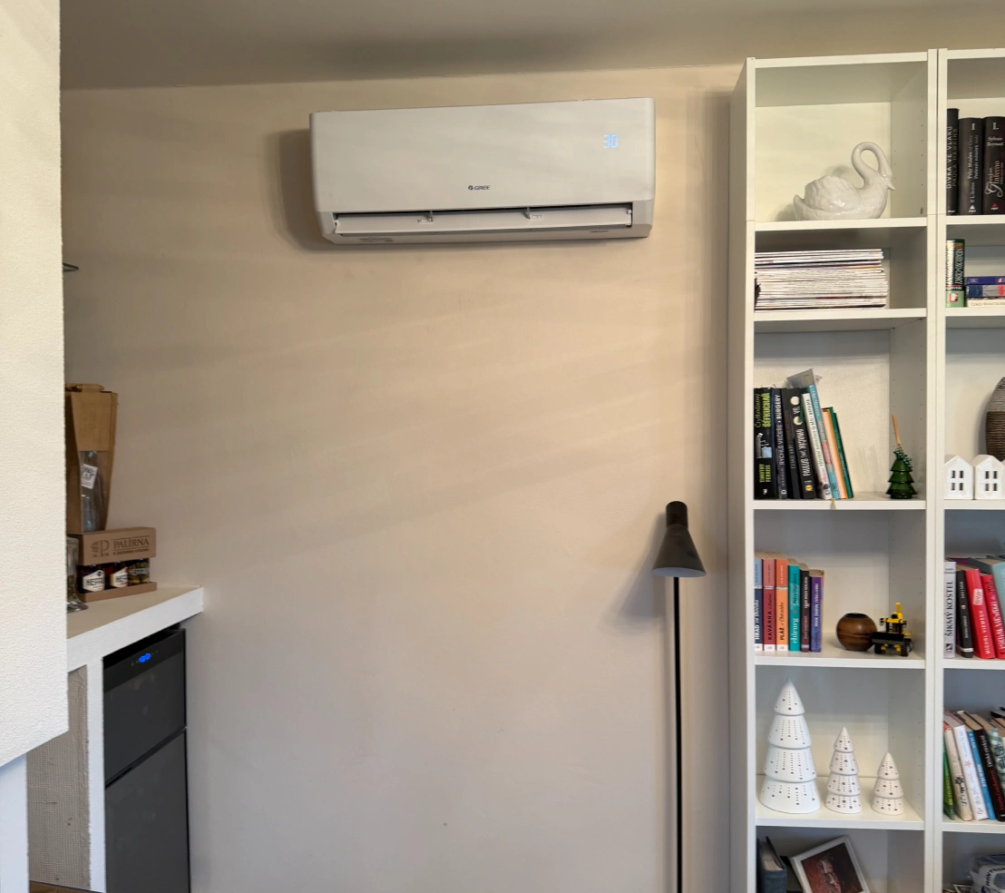 Bílá nástěnná klimatizace Gree s displejem ukazujícím 30 °C vedle knihovny