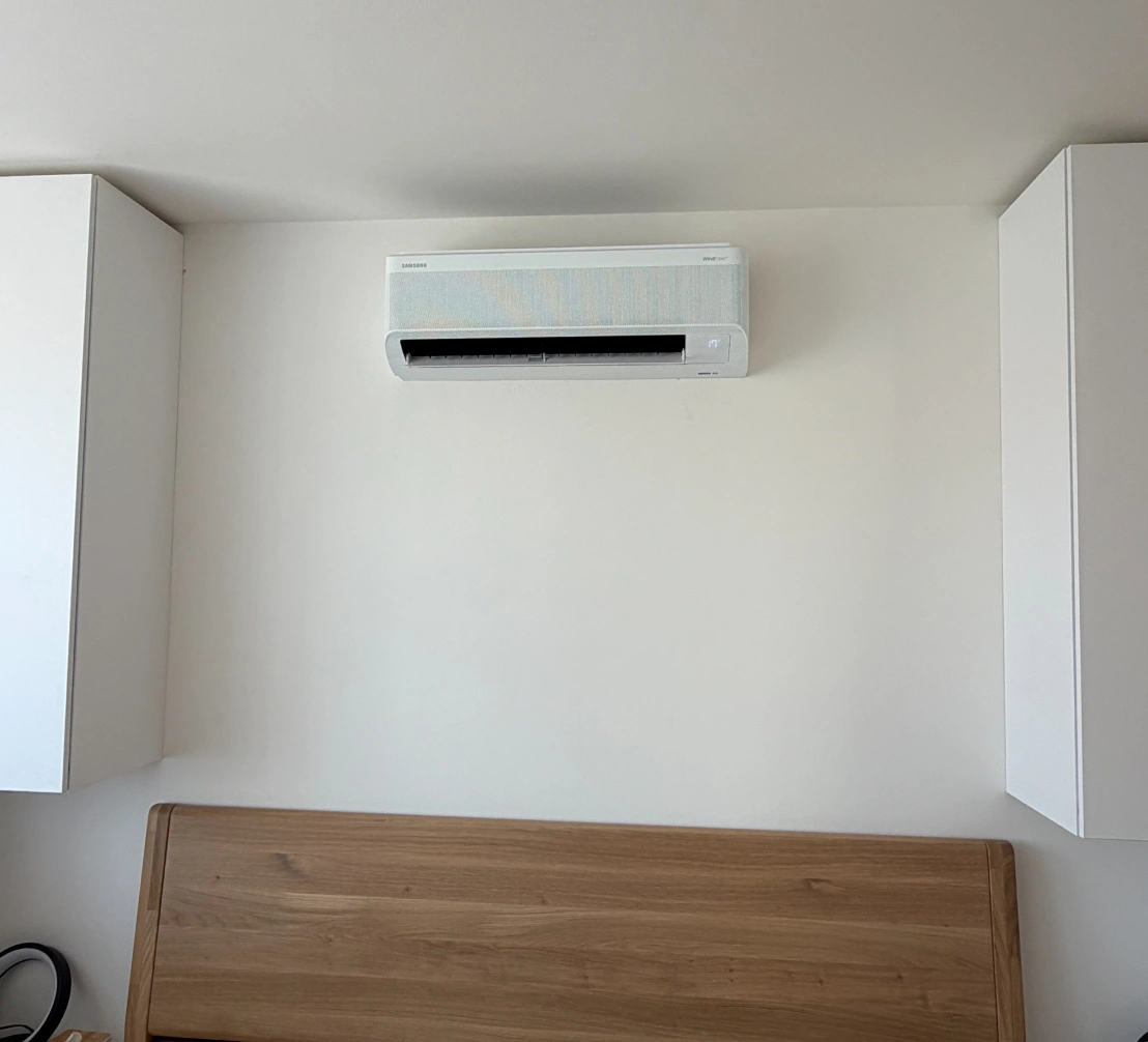 Bílá nástěnná klimatizace Samsung WindFree nad čelem dřevěné postele v ložnici